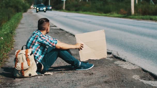 Il giovane si siede a strada in campagna con piatto di cartone in mani. Autostop — Video Stock