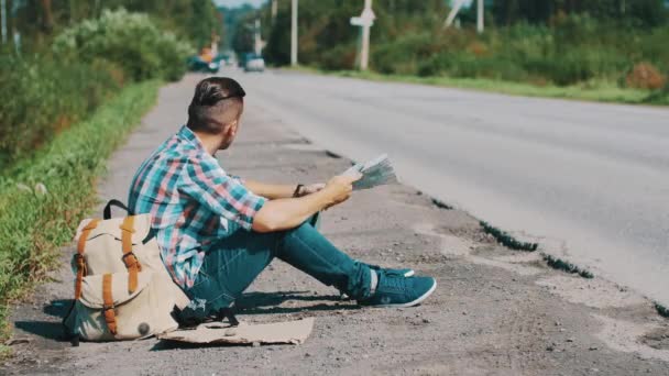 O homem senta-se na estrada no campo. A pedir boleia. À espera. Olhando mapa. Dia ensolarado — Vídeo de Stock