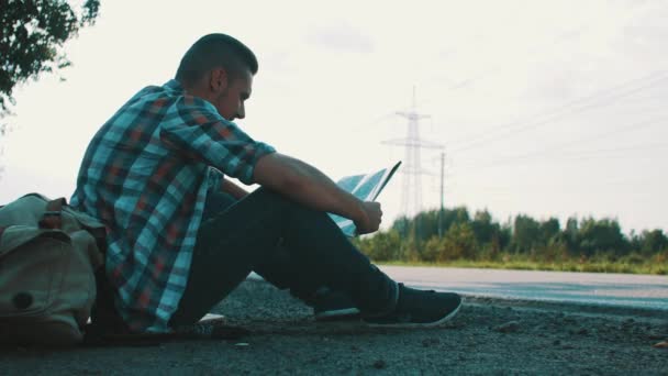 Hombre sentado en la carretera en el campo con el mapa en las manos. Hacer autostop. Esperando. . — Vídeo de stock