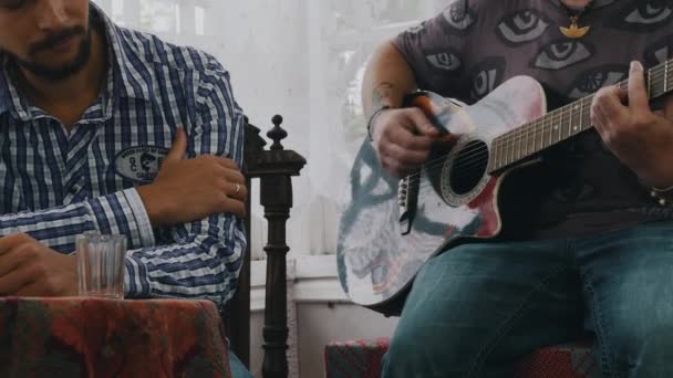 Homme barbu écouter homme jouer de la guitare à table avec des boissons alcoolisées. Maison de campagne — Video