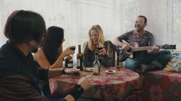 Los amigos levantan la copa con las bebidas en la casa de campo a la mesa. Hombre tocando guitarra — Vídeo de stock