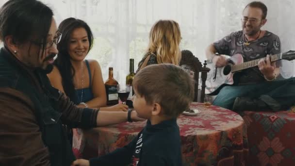 Gli amici in casa di campagna a tavola bevono alcolici. Un uomo che suona la chitarra. Il bambino è venuto — Video Stock