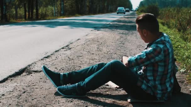 Muž sedí na silnici v krajině s lepenkové desky znak města. Stopování. Slunce — Stock video