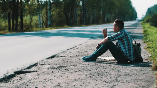 Sad man sit at road in countryside. Hitchhiking. Waiting. Smoking cigarette. — Stock Video