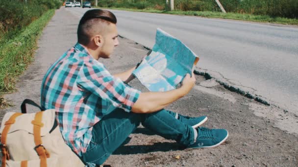 L'homme est assis sur la route à la campagne. L'auto-stop. J'attends de l'aide. Recherche dans la carte . — Video