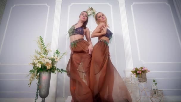 Dvě ženy ve večerních šatech tančí a zpívají ve velkém modrém pokoji s květinami. — Stock video