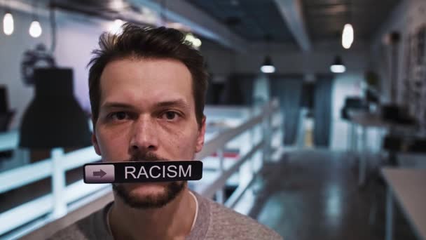 Мужчина смотрит в камеру и держит во рту табличку с надписью RACISM — стоковое видео