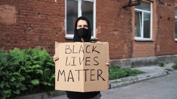 Человек в маске стоит с картонным плакатом в руках - ЧЕРНЫЙ ЖИЗНЬ имеет значение — стоковое видео