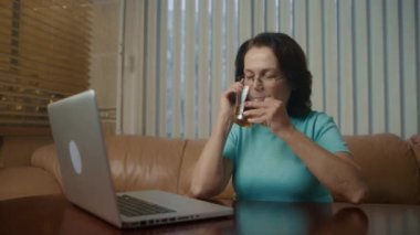 Gözlüklü yaşlı kadın laptopta çalışıyor, neşeli bir şekilde akıllı telefondan konuşuyor. 