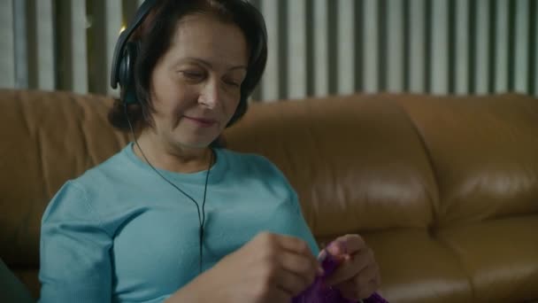 Kvinna stickar halsduk med ekrar, lyssnar på musik med hörlurar och danser — Stockvideo