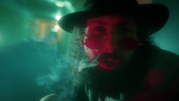 Mannen i svart hatt och glasögon röker en cigarett och tittar in i kameran. — Stockvideo