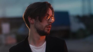 Gözlüklü, şaşkın ve perişan bir adam, sokakta yanan bir arabanın yanında duruyor.