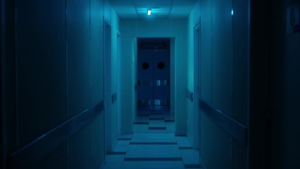 コートを着た男が病院の濃い青の廊下を歩いている — ストック動画