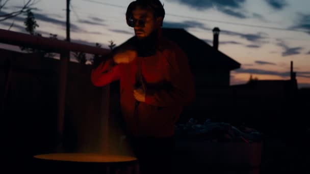 Gözlüklü bir adam ve yüzünde yanan bir çöp tenekesinin yanında güneşlenen kırmızı bir ceket.. — Stok video