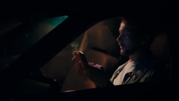 Mannen sitter i bilen på natten, röker en cigarett och pratar i telefon. — Stockvideo