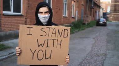 Maskeli adam elinde karton posterle duruyor.