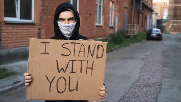 Чоловік у масці стоїть з картонним плакатом в руках - I STAND WITH You — стокове відео
