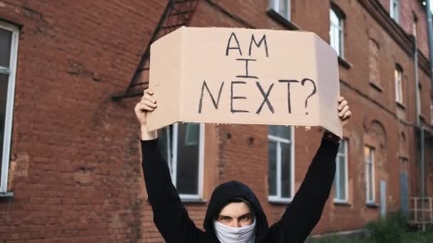 Mężczyzna w masce stoi z tekturowym plakatem w rękach - AM I NEXT — Wideo stockowe