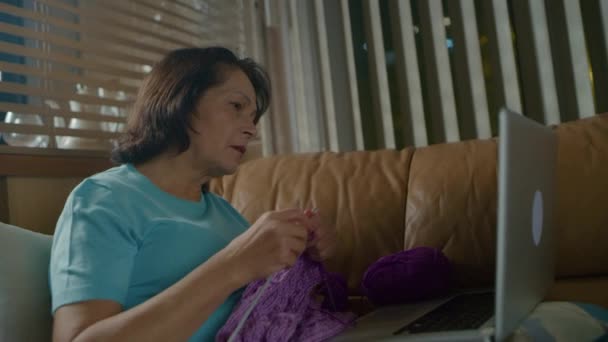 Starsza kaukaska kobieta dzierga fioletowy szalik i używa laptopa do wideokonferencji. — Wideo stockowe