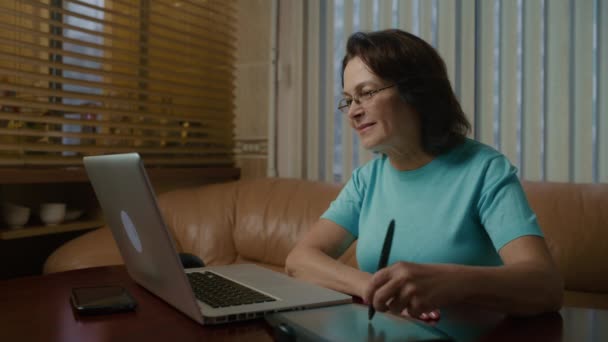 Una donna lavora in un appartamento con un portatile. Disegna immagini utilizzando un tablet — Video Stock