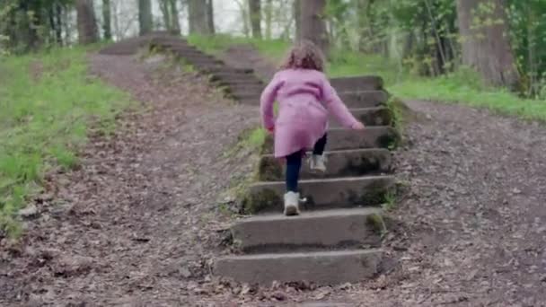 Liten flicka i rock körs längs trappan och gräs i en park med träd — Stockvideo