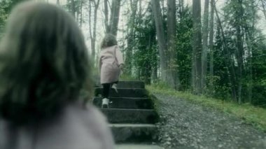 Palto giymiş küçük bir kız merdivenler boyunca koşar ve ağaçlı bir parkta çimenler