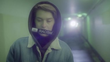 Kapüşonlu ve kot ceketli bir kadın üzerinde RACISM yazan bir tabela taşıyor.