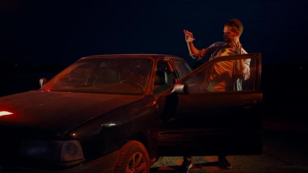 Мужчина ночью танцует, прыгает и машет руками возле машины с открытой дверью. — стоковое видео