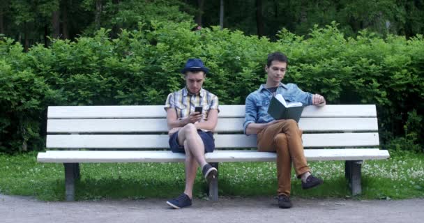 Two guys are sitting on the bench. One is reading a book, the other looking at phone — Stock Video