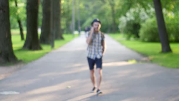 Un chico camina en cámara y habla por teléfono enfocado en salir. Parque verde de verano. Sonríe — Vídeo de stock