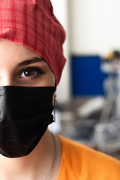 Close-up of half of the face of an attractive young doctor wearing a black surgical mask, a red surgical cap and an orange uniform, prepared to enter the operating room, With a detailed view of a large eye with a brown pupil and a defocused backgroun