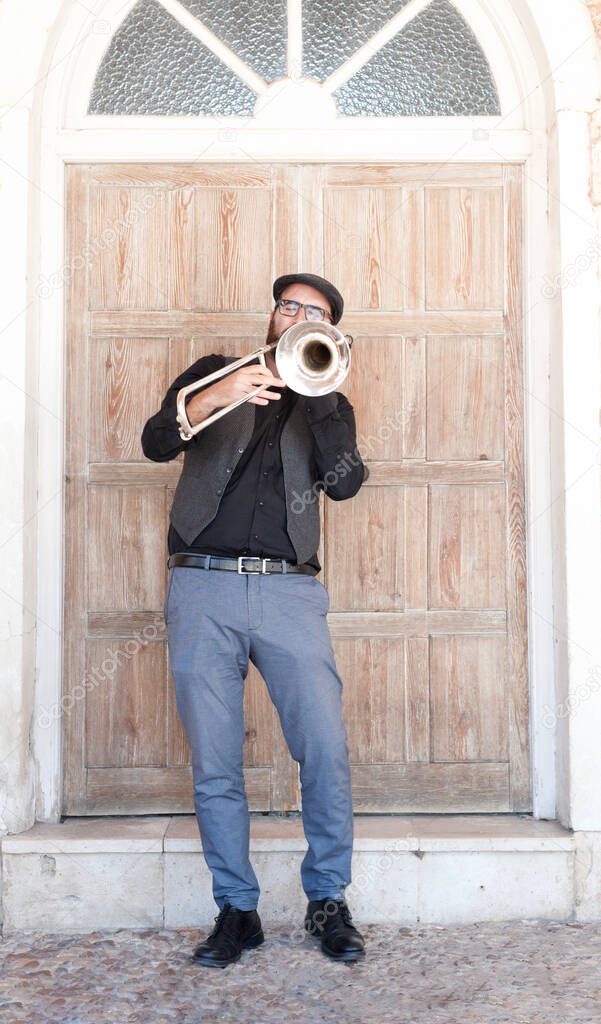 Jazz musician with glasses and beard playing trombone in the street standing. Dressed in an English cap, a jacket, blue trousers. Behind is a wooden doorway behind a step and a glass mezzanine. On the ground a cobblestone street. 
