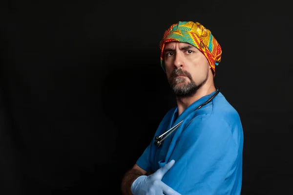 Doctor on the side looking straight ahead with a serious look on his face and his arms crossed. He is wearing a blue scrub shirt, blue surgical gloves, a stethoscope around his neck and an ethnic surgical cap.