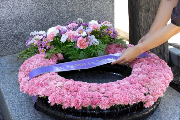 Kadının elleri mezarlıktaki bir mezara çelenk takıyor. Üzerinde kurdele olan gri mermer bir mezar ve pembe karanfil çelengi. 