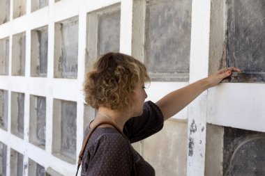 Sarışın ve kıvırcık saçlı bir kadın. Ciddi ve kederli bir jestle mezarı ziyaret ediyor. Ya da bir akrabasını ziyaret ediyor ve içinde bir taş bırakıyor. Ruhlar Günü ya da Azizler Günü Konsepti.