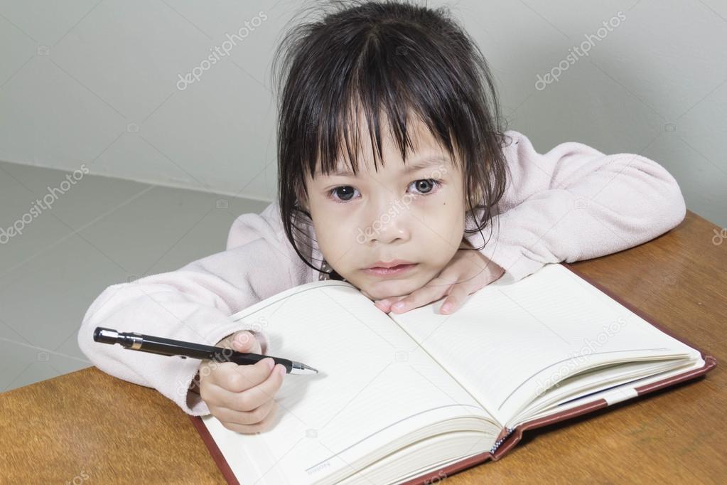 Asian girl kid hard at work in the room sitting with his head on her hand writing notes on book of white paper
