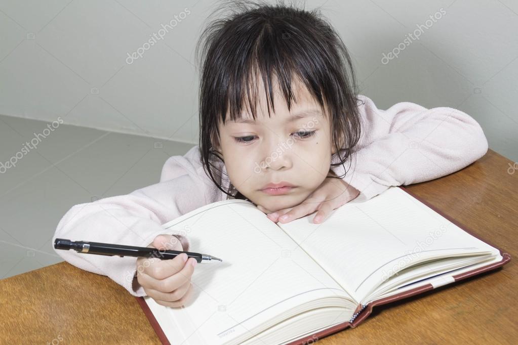 Asian girl kid hard at work in the room sitting with his head on her hand writing notes on book of white paper