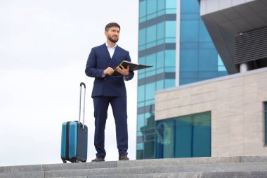 Başarılı bir iş adamı, elinde belgelerle bir ofis binasının arka planındaki basamaklarda duruyor.