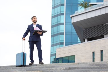 Mutlu iş adamı, elinde iş projesi belgeleriyle ofis binasının arka planındaki basamaklarda seyahat valiziyle ayakta duruyor.