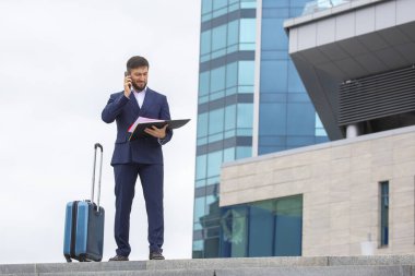 Kendine güvenen bir iş adamı, bir ofis binasının arka planında merdivenlerde duran bir bavulla duruyor.