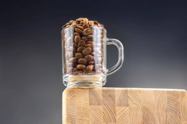 Kavrulmuş kahve çekirdekleri ile dolu şeffaf bir cam bardak koyu mavi arka planda, ahşap bir bloğun kenarında oturur. Minimalist bir natürmort kahve konsepti.