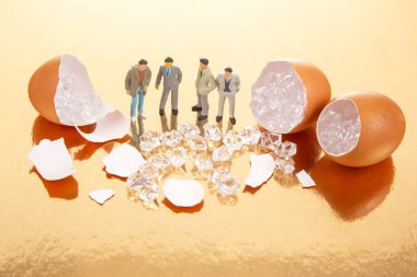 Minyatür heykelcikler, altın bir yüzeyde parıldayan kristallerle dolu kırık yumurta kabuklarının arasında, gerçeküstü bir düzende keşfi, zenginliği veya gizli değeri simgeleyen,