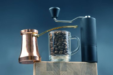 Bakır bir mağara, kahve çekirdekleri ile dolu bir cam bardak ve koyu arkaplana karşı ahşap bir blokta duran bir el yapımı kahve öğütücüsünün yer aldığı şık bir natürmort..