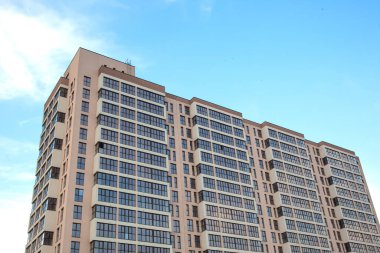 Açık mavi gökyüzünün altında bej bir cephesi ve büyük cam pencereleri olan modern bir gökdelen. Mimari tasarım temiz hatlara ve birden fazla balkona sahiptir.