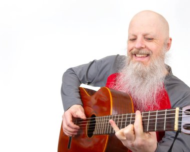 Saçsız ve uzun beyaz sakallı yaşlı bir adam gitar çalıyor, kırmızı ve gri atletik bir bluz giyiyor, beyaz bir arka planda fotoğrafı çekiliyor, bir müzik hobisini vurguluyor.