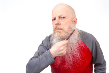 Kalın gri sakallı kıdemli kel adam kırmızı bir tarak kullanıyor, kırmızı ve gri bir spor gömlek giyiyor, beyaz bir arka planda izole edilmiş, ilginç bir kişisel bakım sahnesi yakalıyor.