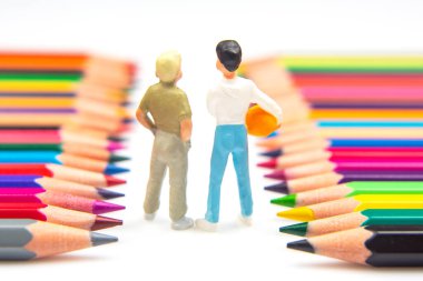 Elinde bir alet tutan küçük bir işçi heykeli, doğumda minimalist beyaz bir arkaplan üzerindeki yaratıcılığı sembolize eden canlı renkli kalemler arasında geziniyor.