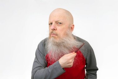 Saçsız ve uzun beyaz sakallı yaşlı bir adam sakalını kırmızı bir tarakla tarıyor, kırmızı ve gri atletik bir bluz giyiyor, komik bir portre için temiz beyaz bir zemin üzerinde fotoğraf çekiliyor.