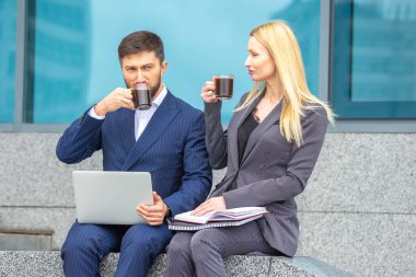 İş adamları ve kadınlar ellerinde belgeler ve dizüstü bilgisayarla bir iş binasının merdivenlerinde oturuyor kahve içiyor ve iş planlarını tartışıyorlar.