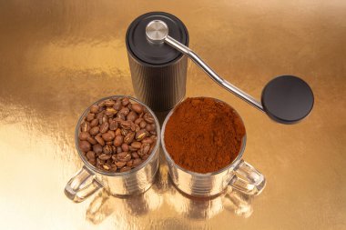 Bir el yapımı kahve öğütücüsü, biri kavrulmuş kahve çekirdekleri, diğeri taze öğütülmüş, altın bir yüzeyde, iki cam bardağın arasında oturur ve kahve öğütme işlemini gösterir.